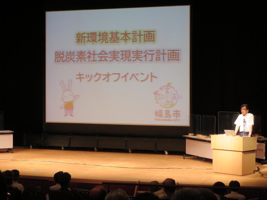 ステージ上でスクリーンに写し出される資料をもとに講演をする市長と、多くの参加者の方々の写真