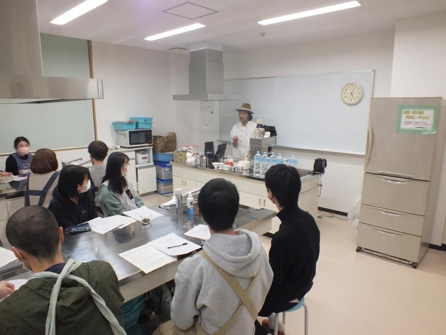 ホワイトボードの前に講師の先生が立ち、向かい合わせの机に参加者の人たちが座り説明を聞いている写真
