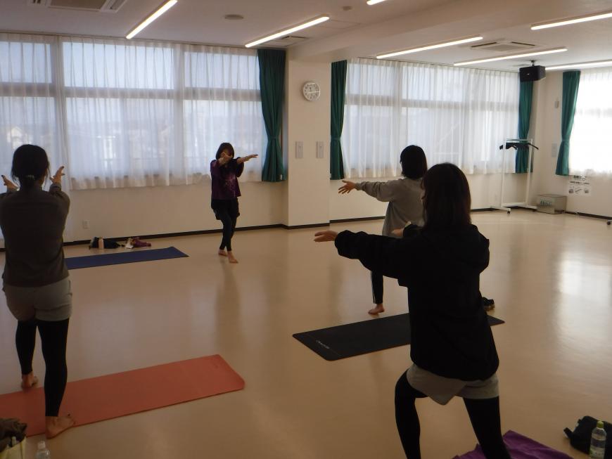 窓側に講師の女性が右足を曲げて前に出し両手を前に出しているポーズを参加者の人もマットの上で同じポーズをしている写真