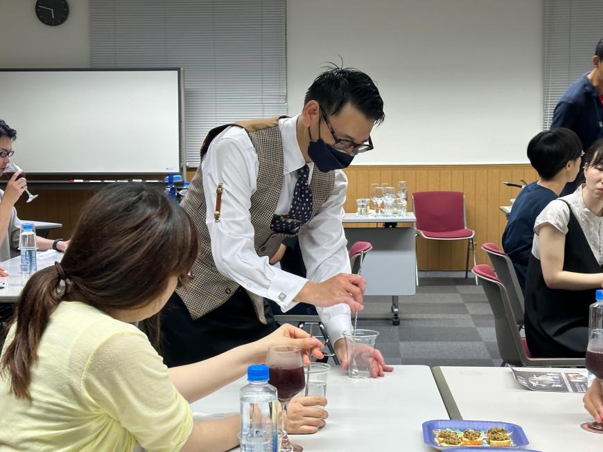 講師の先生が女性の横でバースプーンを使いながらグラスの混ぜ方を指導している写真