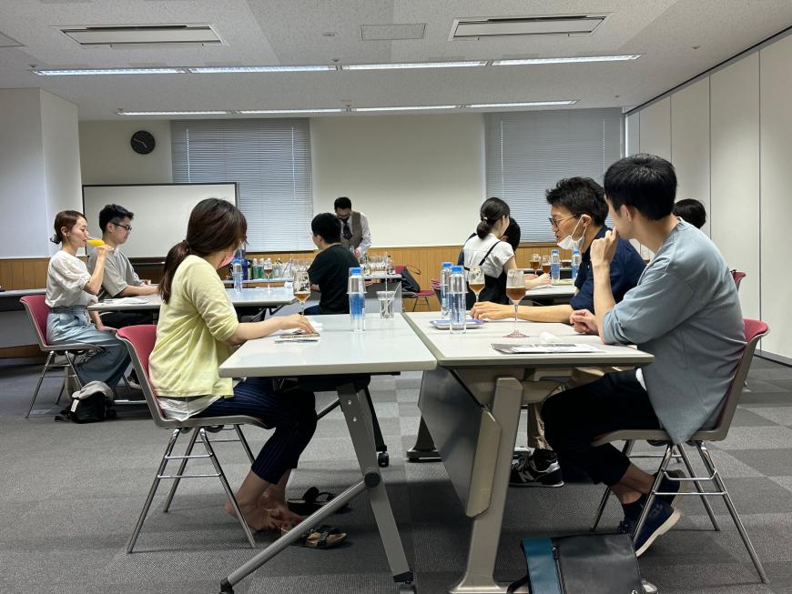 参加者が3名ずつ座った3つのグループがあり、テーブルにはボトルとグラス、正面に講師の先生がいる室内写真