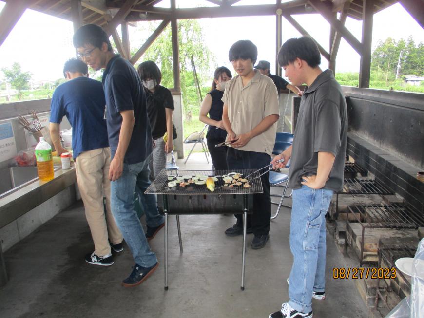 屋外炊事場で男性2名がトングでお肉や野菜などを網で焼きながらバーベキューを楽しんでいる男女7名の写真