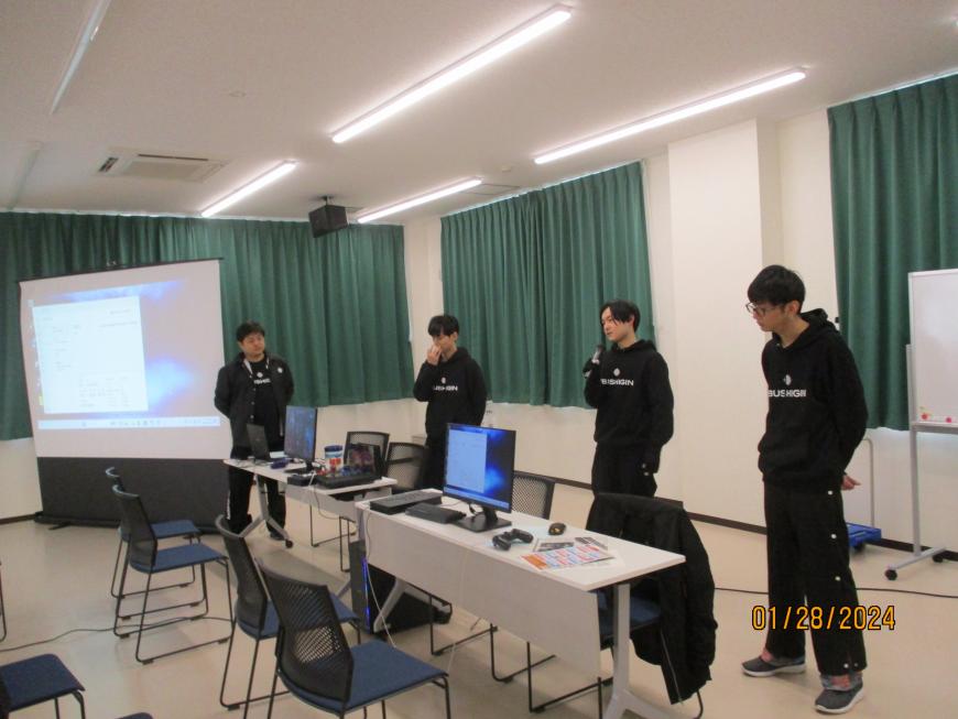 4名の男性が並び右から2人目の男性がマイクを持っている写真