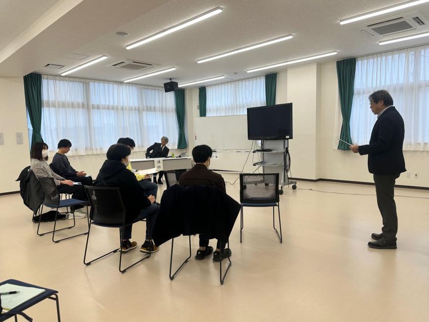 5人の参加者が椅子に座りながら右側に立つ男性の説明を聞いている写真