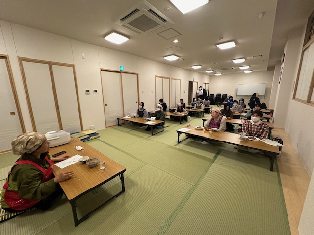 和室の長机に料理が置かれ、2名ずつ参加者が座り講師の先生の説明を聞いている写真