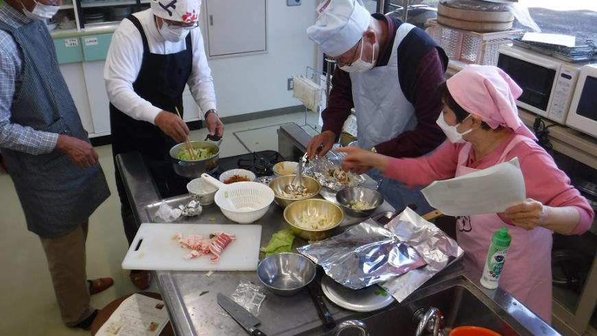 男性3名が各担当で料理をしている横で女性が指導をしている料理教室での写真