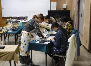 中央の机にテクスチャ―アートで使用する様々な材料が準備され、長机に座っている参加者の女性に先生が指導をしている写真