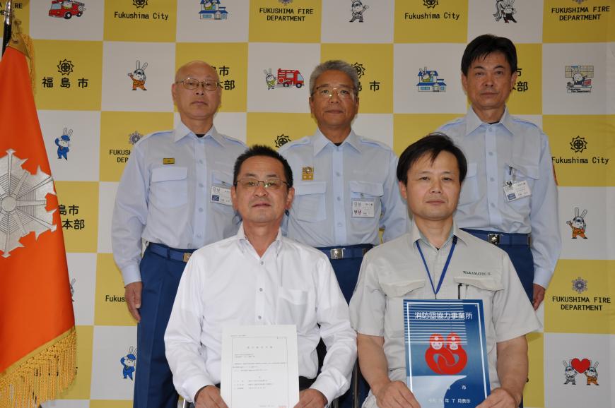 バックボードの前で前列に2人が座り消防団協力事業所表示証を持ち、後ろに3人が立って記念撮影している写真