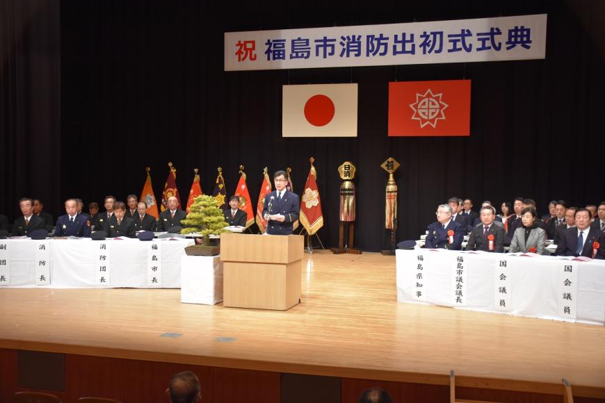 「福島市消防出初式典」と書かれた横断幕と、国旗と市旗が掲げられたステージ上に関係者が着席し、檀上で市長が挨拶をしている写真