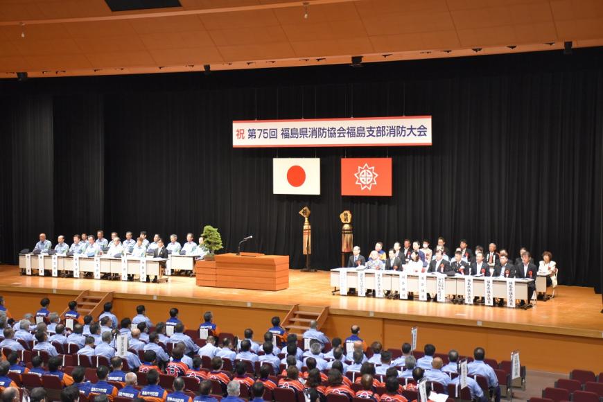 舞台に「第75回福島県消防協会福島支部消防大会」と横断幕が掲示され、舞台上には関係者が着席し、参加者の皆さんが会場の客席に着席している会場の様子を写した写真