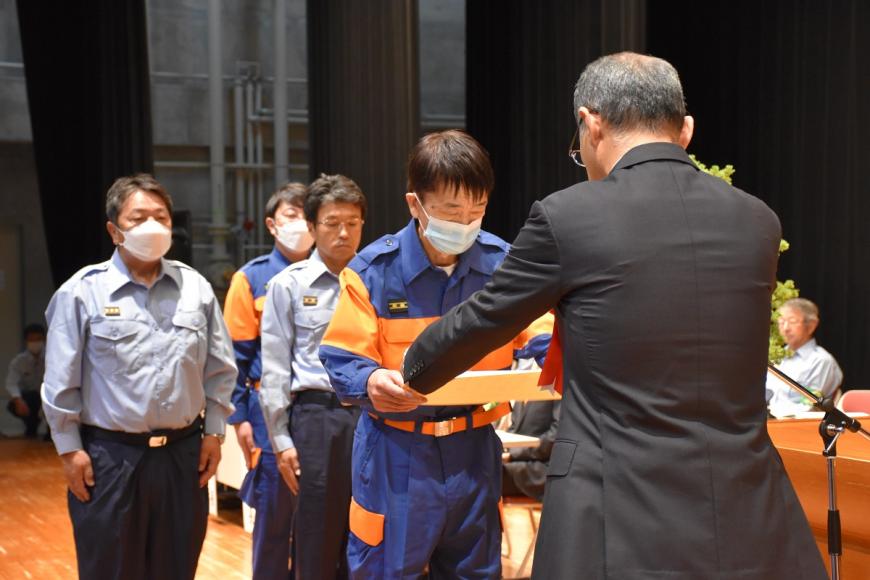 関係者が後ろに数名立っており活動服を着た男性が福島県消防表彰を受け取っている様子の写真