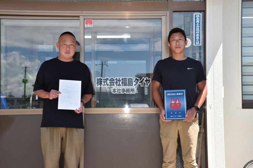 株式会社福島タイヤの建物の前で男性2人が消防団協力事業所表示証を持っている写真