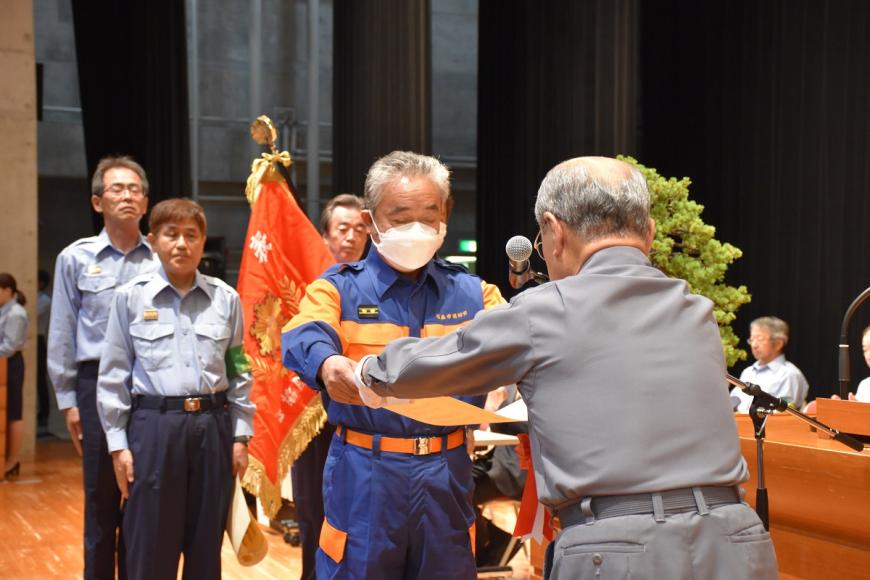 関係者が後ろに数名立っており活動服を着た男性が福島県消防協会定例表彰を受け取っている様子の写真