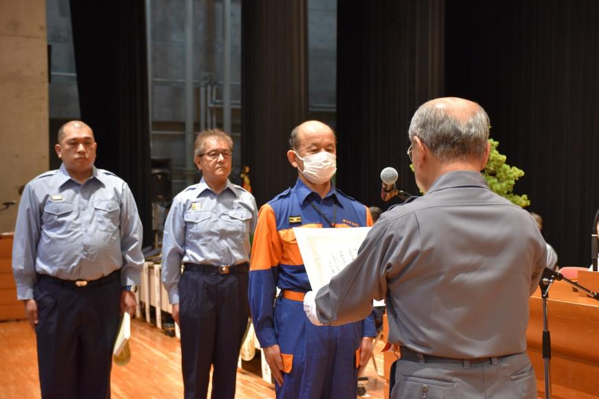 関係者が後ろに数名立っており活動服を着た男性が福島県消防協会定例表彰を受け取っている様子の写真