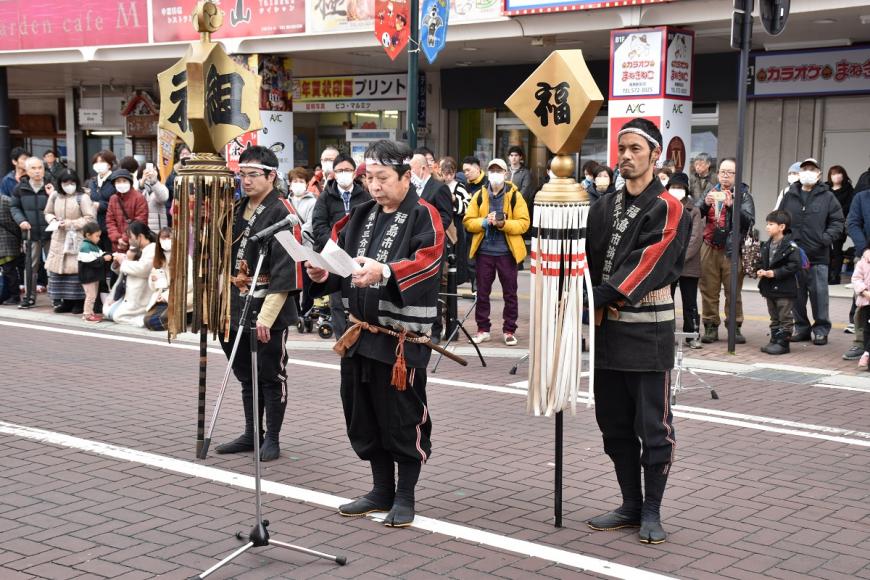 黒い法被を着た2名の男性が纏を持ち並んでいる中央の男性が決意表明を述べている様子の写真