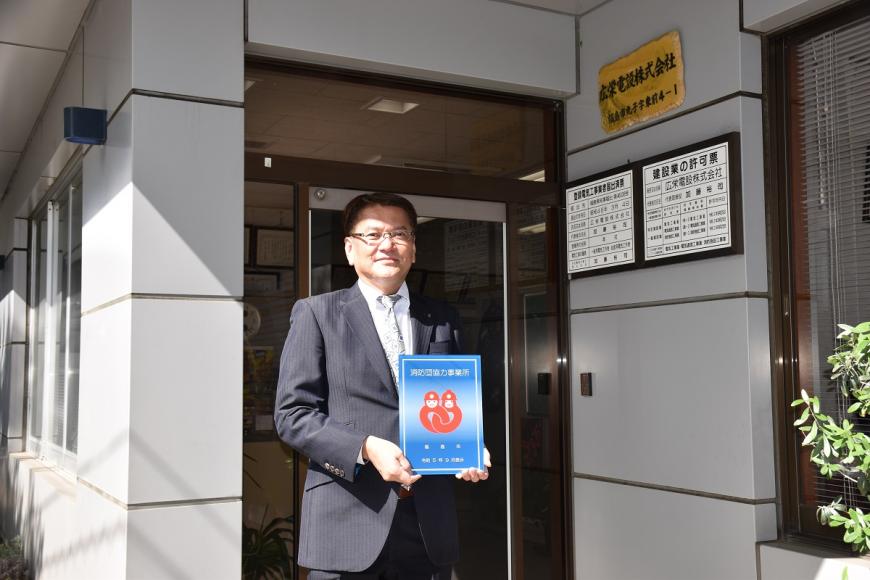 広栄電設株式会社の建物の前で男性が消防団協力事業所表示証を持っている写真