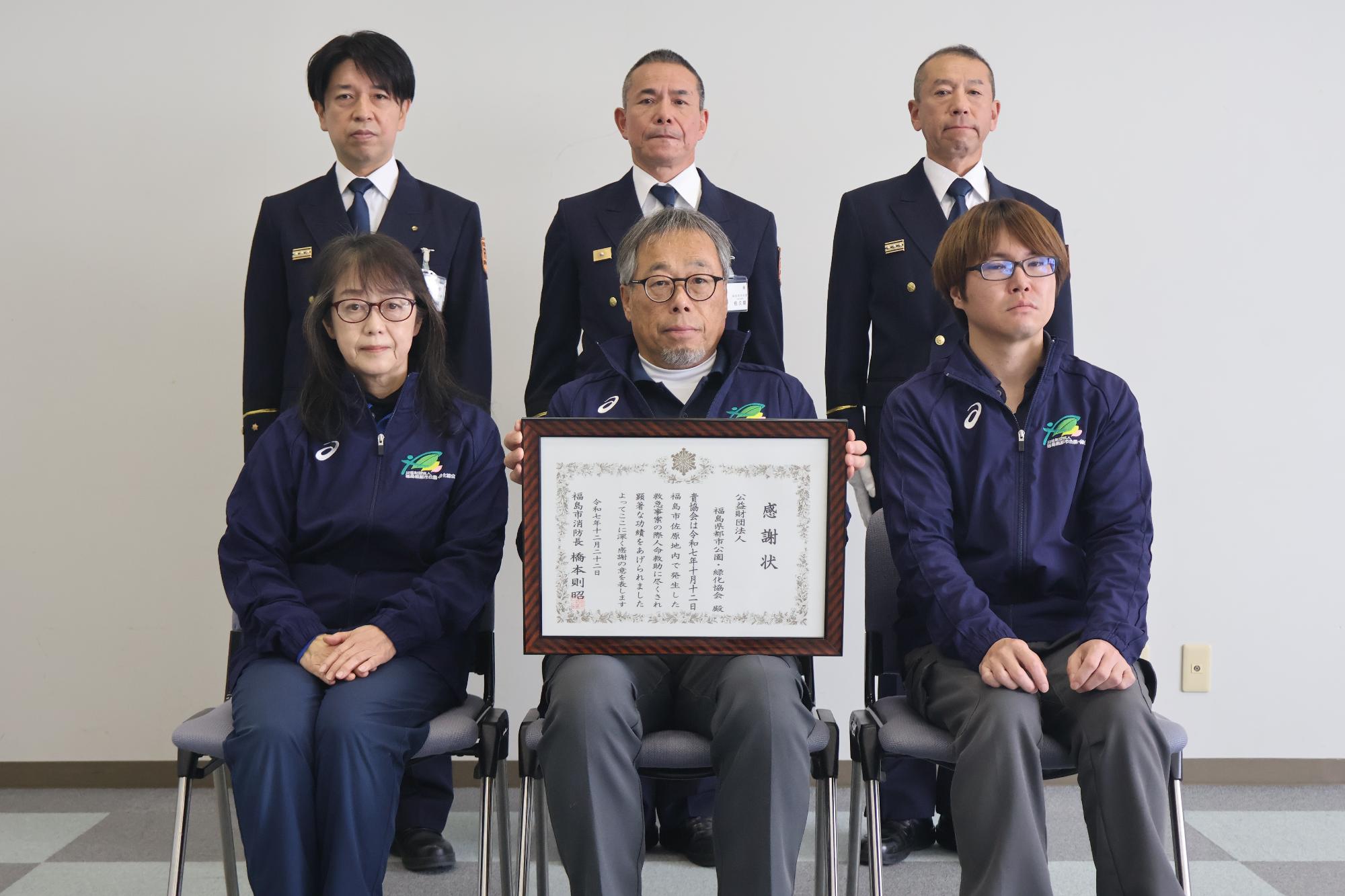 受章者3名が椅子に座り、中央で代表受章者が感謝状を持って記念撮影した写真