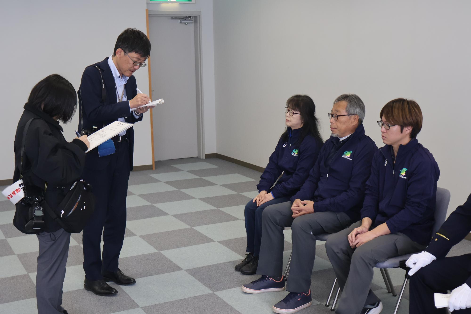 受章者3名が椅子に座り、報道機関の取材を受けている写真