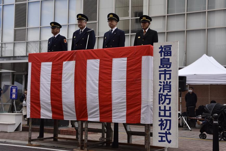 紅白の横断幕が貼られた台の上に、帽子を被り制服姿の市長を含め4名の男性が横一列に並んでいる写真