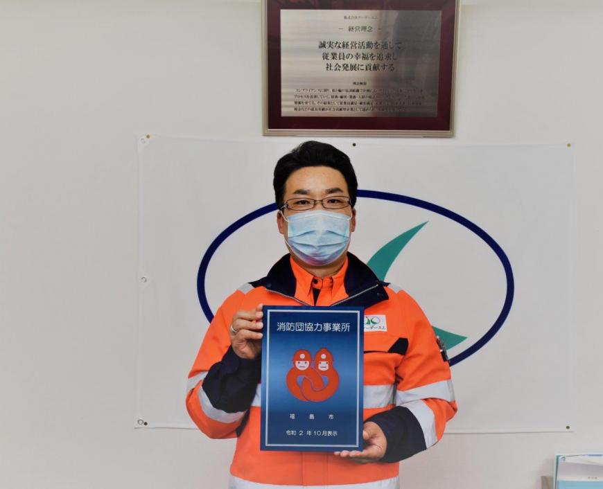 オレンジの制服を着た男性が防団協力事業所表示証を持っている写真