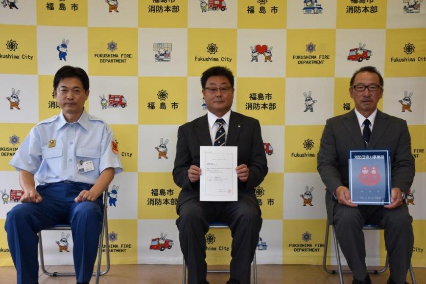 バックボードの前で3人が椅子に座り2人が消防団協力事業所表示証を持って記念撮影している写真