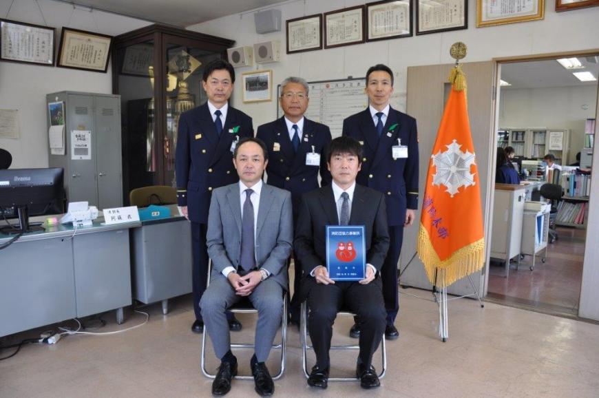 前列に2人が座り、1人が消防団協力事業所表示証を持ち、後ろに3名が立っている記念写真