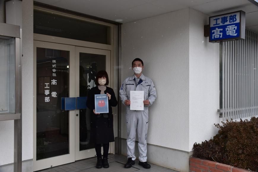 株式会社高電の建物の前で女性と男性が消防団協力事業所表示証を持っている写真