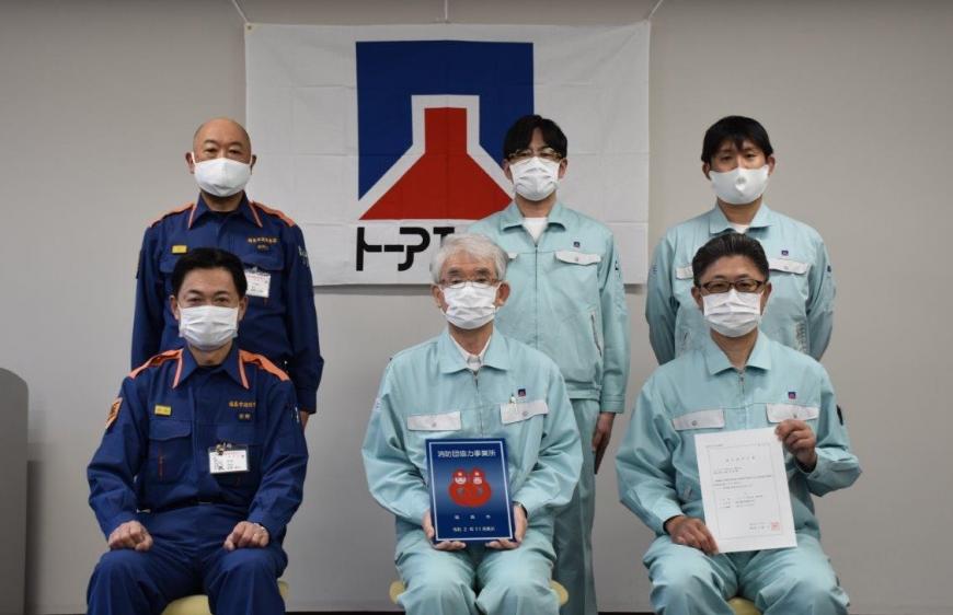 前列に椅子に座り3人の2人が防団協力事業所表示証を持って、後ろの3人が立って並んで記念撮影している写真