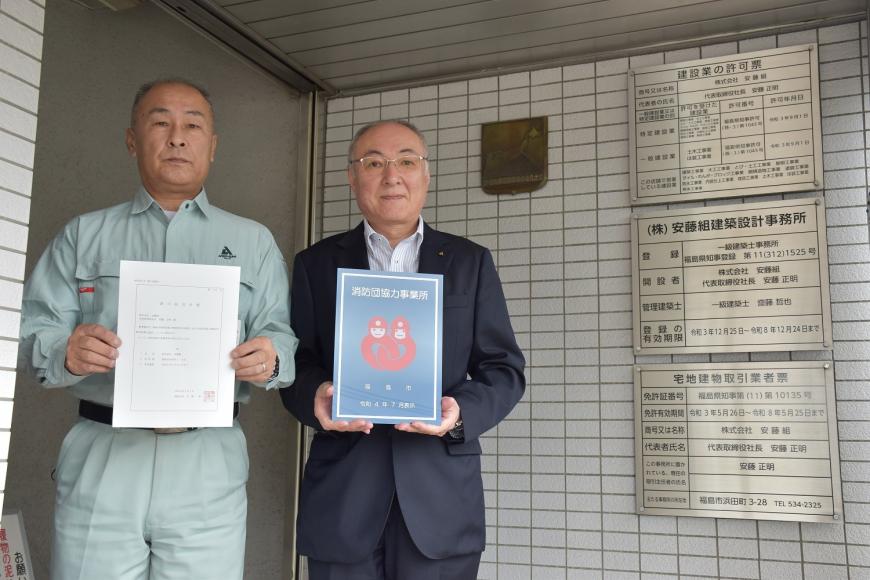 株式会社安藤組の建物の前で男性2人が消防団協力事業所表示証を持っている写真