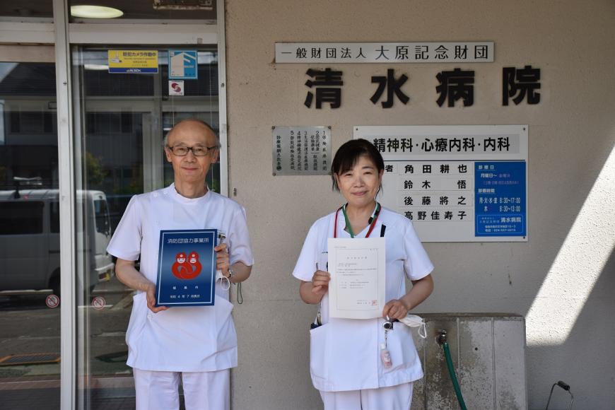 一般財団法人大原記念財団清水病院の建物の前で白衣を着た男性と女性が消防団協力事業所表示証を持っている写真