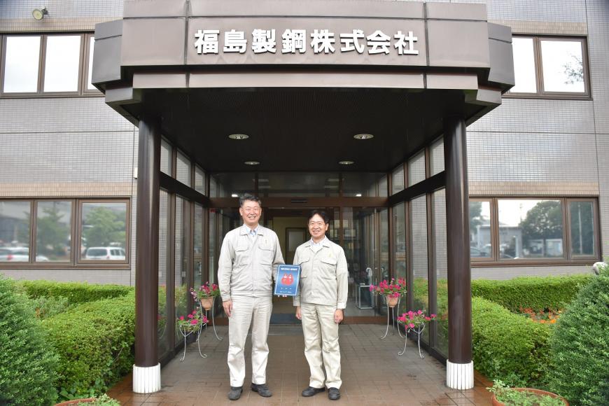 福島製銅株式会社の建物の前で男性2人が消防団協力事業所表示証を持っている写真