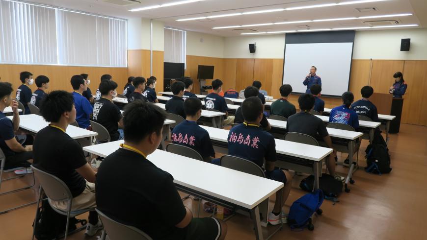 スクリーンの前でマイクを持って話をする職員の話を聞く参加者の写真