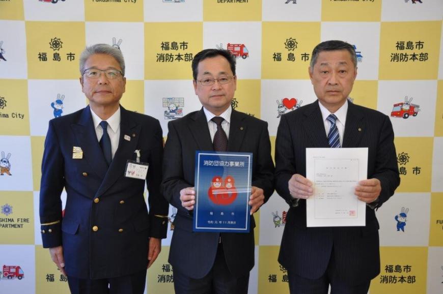 バックボードの前で制服姿の2人が消防団協力事業所表示証を持ち、3人で記念撮影している写真