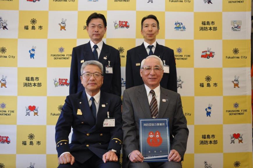 バックボードの前で前列に2人が座り1人が消防団協力事業所表示証を持ち、後ろに2人が立って記念撮影している写真