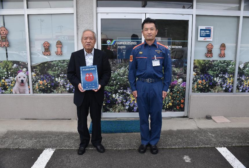 八巻興業株式会社の建物の前で社員2人が消防団協力事業所表示証を持っている写真