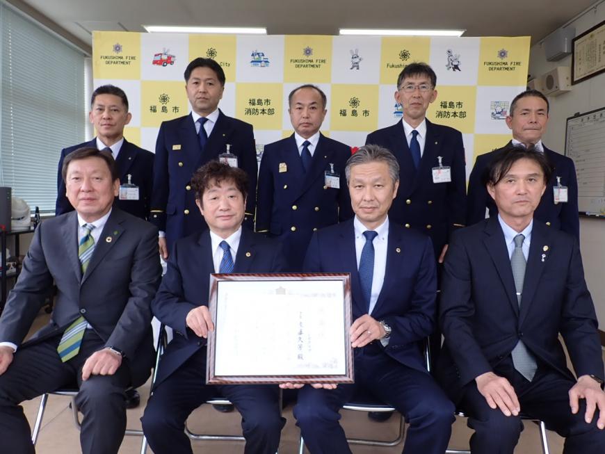 福島市と福島市消防本部のバックボードを背景に関係者が2列に並び前列の2名が一緒に贈呈の書状を持っている記念撮影の写真