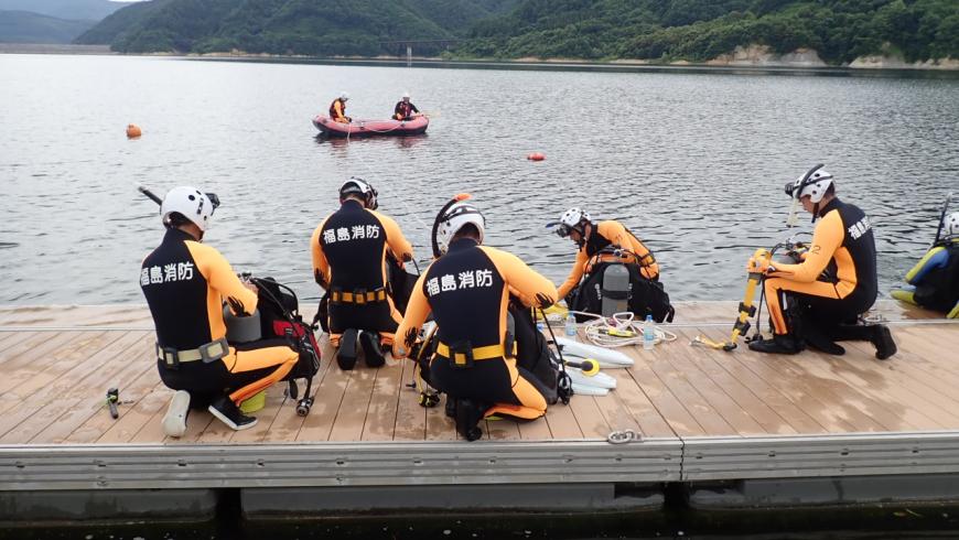水辺の近くで「福島消防」とかかれたウェットスーツを着用した数名の隊員が装備を確認している水難救助隊訓練の様子の写真